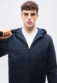 Νέος άνδρας που φοράει σκούρα μπλε ζακέτα με φερμουάρ πάνω από λευκό πουκάμισο με γιακά, κρατώντας ένα λουρί πάνω από τον δεξί του ώμο μπροστά σε απλό φόντο.