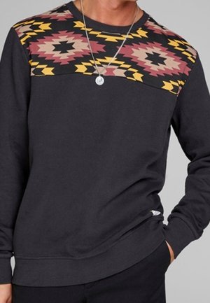 Czarna bluza z geometrycznym, plemiennym wzorem w żółtym, różowym i czarnym na klatce piersiowej i ramionach, noszona z srebrnym naszyjnikiem z zawieszką.