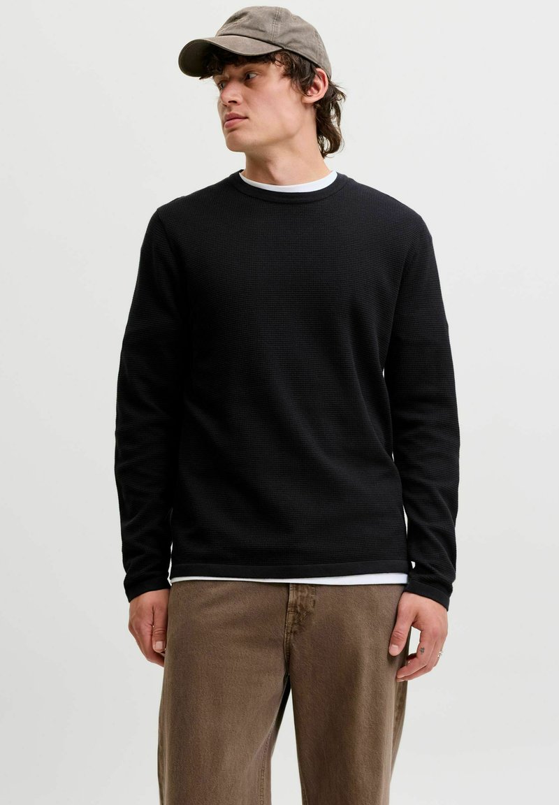Jeune homme portant une chemise noire à manches longues, un pantalon marron et une casquette grise regardant sur le côté sur un fond clair uni.