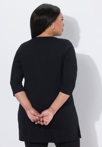 Black tunic with three-quarter sleeves, made of smooth fabric, featuring a rounded neckline and side slits for ease of movement.