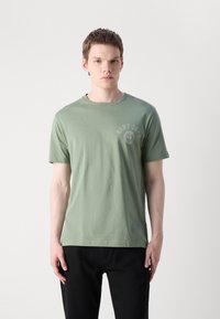 Šviesiai žalia medvilninė marškinėlių T-shirt su trumpomis rankovėmis, apvaliu iškirpimu ir apvaliu "GANT 1949" logotipo detale kairėje krūtinės pusėje.