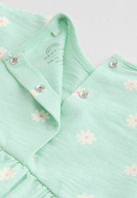Otroško oblačilo v metini zeleni barvi z belimi in roza potiski marjetic, z srebrnimi sponkami ob vratnem izrezu in prsnem predelu.
