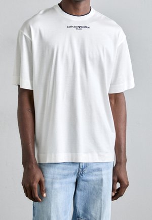 Homme portant un t-shirt blanc ample Emporio Armani Milano et un jean bleu clair, debout devant un fond neutre.