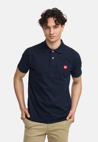 Marineblå polo skjorte laget av bomull. Har krage, tre-knapp placket og en liten rød logo på venstre bryst. Lettvektig stoff.