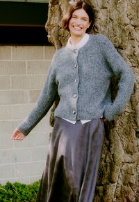 Cardigan grigio a maglia con colletto tondo, cinque pulsanti frontali e texture a coste, abbinato a una gonna in raso scuro. Il modello si appoggia a un albero.