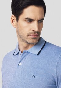Jasnoniebieska koszulka polo z teksturowanej dzianiny, kontrastujące ciemne akcenty na kołnierzyku oraz małe czarne logo wyszyte na piersi.