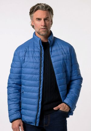 Blå pufferjakke med horisontalt quiltet mønster, høj krave og lynlåslukning. Båret over en mørk striktrøje og denim jeans.