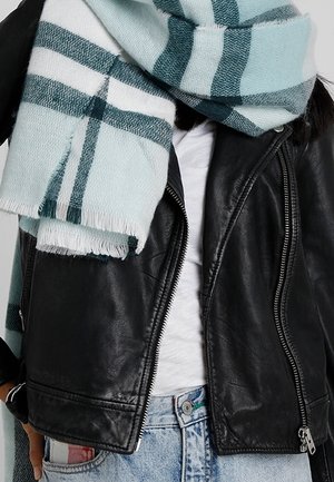 Personne portant une écharpe à rayures bleu clair et vert foncé, une veste en cuir noire zippée, une chemise blanche et un jean bleu avec un détail rouge.