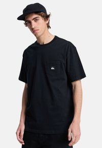 Sort bomulds T-shirt med korte ærmer, rund halsudskæring og en frontlomme med et lille logo. Båret med sorte bukser og en matchende kasket.