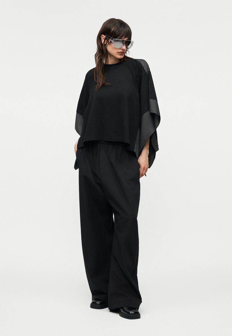 Femme portant un pantalon large noir oversize et un haut ample assorti avec des accents en cuir, accessoirisée de grandes lunettes de soleil teintées.
