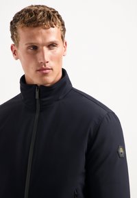 Marine blauwe ritsjack met een hoge kraag, gemaakt van gladde stof. Voorzien van zijzakken en een hexagonaal logo embleem op de schouder.