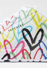 Bijela atletska cipela prekrivena šarenim uzorcima srca i linija, s dizajnom sa vezicama i logotipom "SKECHERS" sa strane.