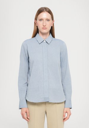 Lyseblå skjorte med krage laget av teksturert stoff, med en frontplacket og lange ermer. Kombinert med beige bukser.