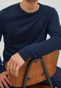 Mężczyzna w granatowym swetrze z długim rękawem siedzący tyłem na drewnianym krześle z metalową ramą, opierający ramię o oparcie krzesła.