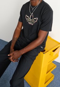 Camiseta negra con un diseño gráfico multicolor en el pecho, combinada con pantalones negros. Sentado en una unidad de almacenamiento amarilla con una superficie texturizada.