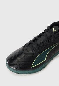Zapatilla deportiva negra de Puma con suela verde oscura, detalles verdes y cordones negros sobre un fondo claro y liso.