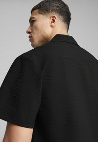 Czarna koszula z krótkim rękawem o fakturowanej tkaninie, z klasycznym kołnierzykiem i widocznymi detalami szycia na plecach.