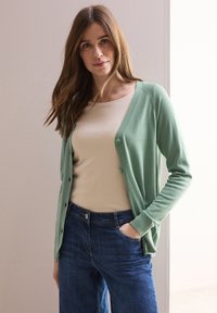 Cardigan verde chiaro con una texture morbida, dotato di una chiusura a bottoni sul davanti, indossato sopra a una maglietta beige e jeans blu. Design semplice, vestibilità casual.