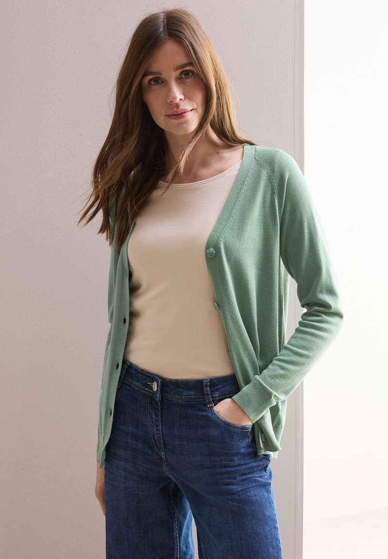 Cardigan verde chiaro con una texture morbida, dotato di una chiusura a bottoni sul davanti, indossato sopra a una maglietta beige e jeans blu. Design semplice, vestibilità casual.
