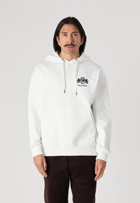 Weißer Hoodie aus weichem Stoff, mit einer Känguru-Tasche vorne und Kordeln. Besticktes Logo auf der Brust. Lange Ärmel.