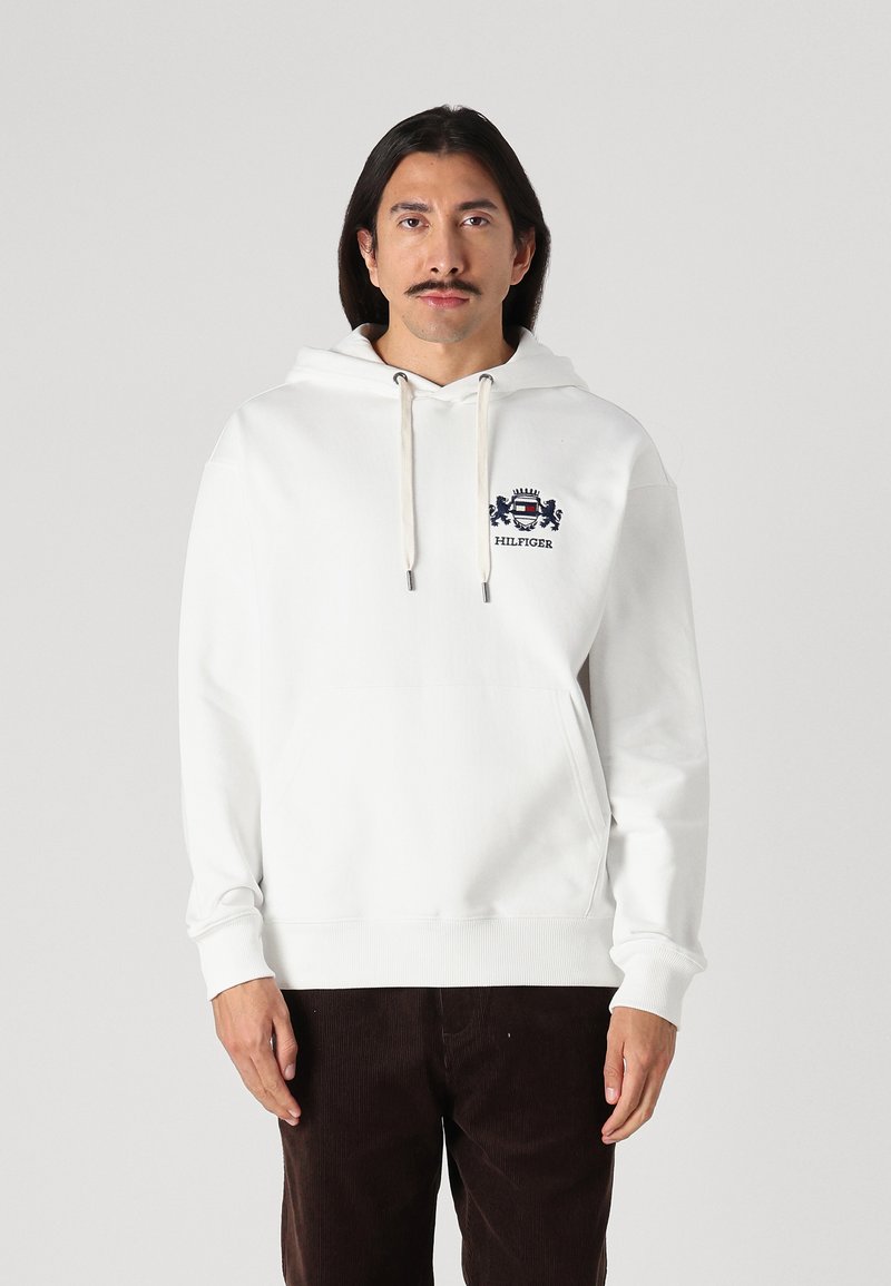 Weißer Hoodie aus weichem Stoff, mit einer Känguru-Tasche vorne und Kordeln. Besticktes Logo auf der Brust. Lange Ärmel.