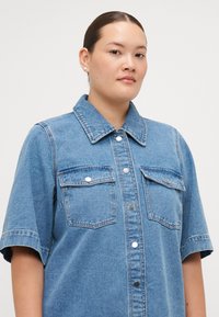 Denim shirt in light blue, featuring a button-up front, two chest pockets, short sleeves, and a structured collar. Smooth texture.