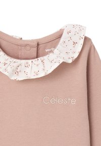 Light pink long-sleeve top with a white floral ruffle collar. Features embroidered text "Céleste" in subtle contrast. Soft fabric texture.