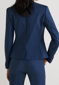 Blazer bleu marine ajusté, doté d'une coupe ajustée, d'épaules structurées, d'une petite fente au dos et d'un tissu lisse et texturé.