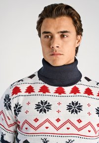 Pull tricoté blanc avec des motifs festifs rouges et noirs, doté d'un col roulé gris foncé et de motifs sur le thème des fêtes.