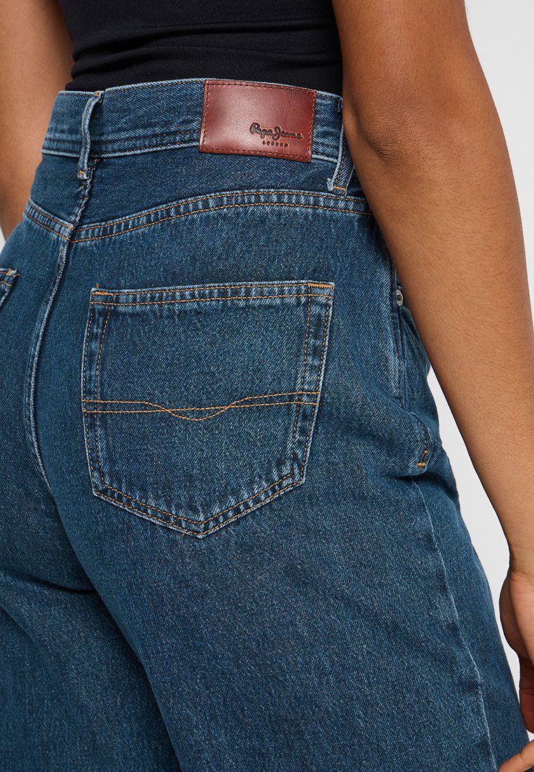 Donkere spijkerbroeken met hoge taille, met een bruine leren achterpatch en een enkele achterzak met oranje stiksels.