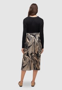 Una donna sta in piedi di spalle indossando un top a maniche lunghe nero e una gonna midi plissettata con un motivo geometrico beige e nero, scarpe slip-on color sabbia.