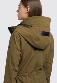 Giacca impermeabile verde oliva con colletto alto, cappuccio regolabile e cordino elastico. Include una patch nera con logo sul retro.