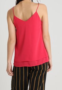 Woman wearing a sleeveless, bright pink V-back top with thin straps and black pants with vertical brown stripes, shown from behind.