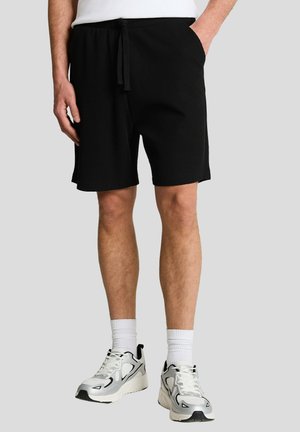 Man wearing black textured shorts with drawstring, white crew socks, and white and black athletic sneakers, standing against gray background.