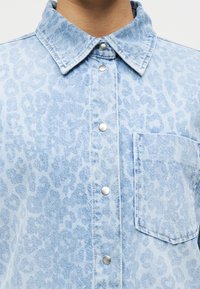 Ανοιχτό μπλε τζιν πουκάμισο με διακριτικό μοτίβο λεοπάρ, εμπρόσθια κουμπώματα, μυτερός γιακάς και τσέπη στο στήθος. Ασημένιες μεταλλικές λεπτομέρειες.