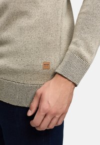 Beige gebreide trui met geribde cuffs en zoom, voorzien van een klein bruin etiket. De textuur lijkt zacht en lichtjes gestructureerd.