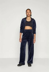 Námornícky zamatový teplákový set s naberanou bundou so zipsom a elastickými manžetami, spolu s širokými nohavicami. Čierny športový podprsenka a nazúvacie topánky dopĺňajú vzhľad.