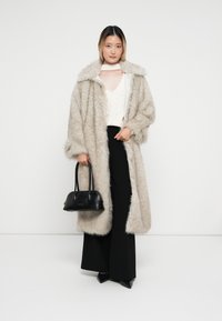 Cappotto in pelliccia sintetica di colore grigio chiaro, con ampio colletto, abbinato a un top in maglia crema, pantaloni neri a gamba larga e una borsa nera. Tessuto liscio.