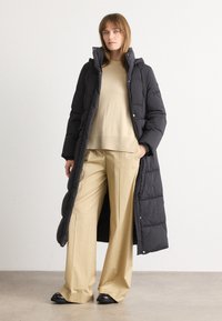 Γυναίκα που φοράει ένα μακρύ μαύρο γούνινο παλτό πάνω από μπεζ πουλόβερ και φαρδιά μπεζ παντελόνια, σStanding against a plain white background.