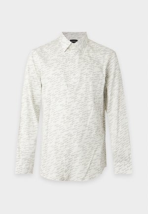 Jeune homme debout portant une chemise blanche à manches longues avec motifs, un jean bleu ample et des chaussures en cuir noires sur un fond uni.