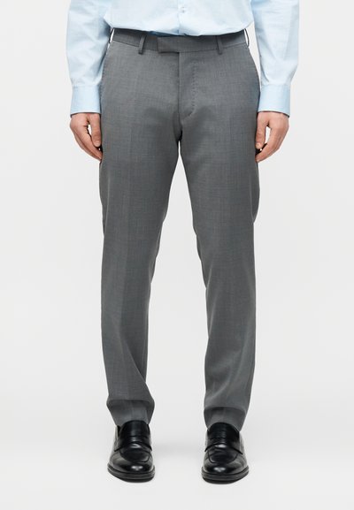 Άντρας που φοράει γκρι ραμμένο στα μέτρα του παντελόνι, ανοιχτό μπλε πουκάμισο με κουμπιά και μαύρα δερμάτινα παπούτσια, στέκεται με χαλαρά τα χέρια στο πλάι.