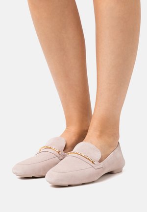 Pieds portant des mocassins en daim rose clair à petits talons avec des accents de chaîne dorée sur un fond blanc.