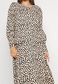 Haut à manches longues en tissu beige avec un motif à pois noirs. Coupe décontractée, encolure arrondie et poignets froncés.