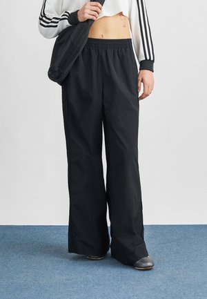 Oseba, ki nosi črne široke hlače, kratko dolgotrajno belo majico s črnimi črtami in drži črno torbo preko rame na modrem preprogi.