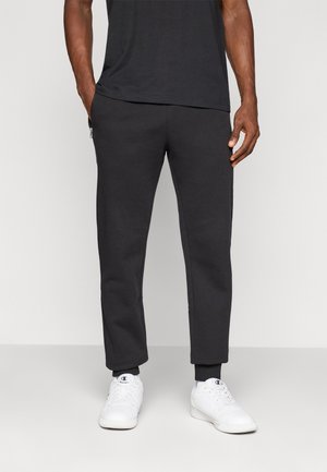 Homme portant un pantalon de jogging noir avec des poches zippées, des baskets montantes blanches et un t-shirt noir, se tenant devant un fond uni.