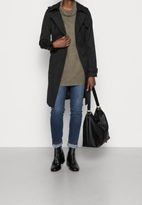 Schwarzer Trenchcoat mit doppelreihigen Knöpfen, über einem olivgrünen Oberteil getragen, kombiniert mit schmalen Jeans und schwarzen Stiefeletten; hält eine schwarze Tasche.