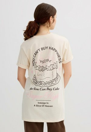 Mujer con cabello castaño recogido, que lleva una camiseta crema con ilustración de pastel y texto "No puedes comprar la felicidad pero puedes comprar pastel" en la parte trasera.