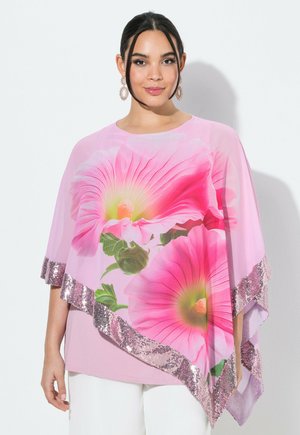 Frau trägt ein pinkes florales Top mit großen Hibiskus-Prints und Paillettenbesatz, kombiniert mit weißen Hosen vor einem schlichten weißen Hintergrund.