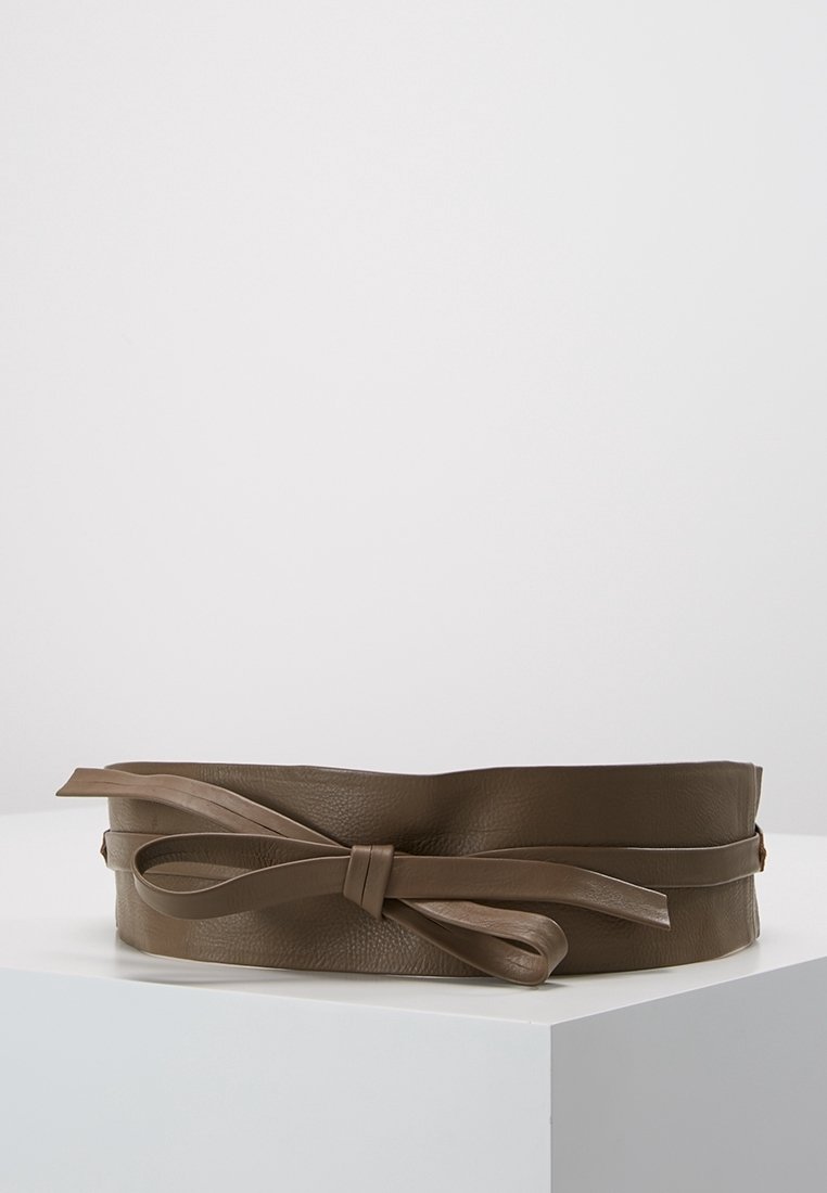 Large ceinture en cuir marron avec un détail en forme de nœud, présentée sur une surface blanche devant un fond gris clair uni.