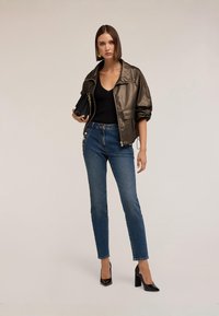 Lesklá zelená bomber bunda olivové barvy přes černý žebrovaný top, doplněná modrými džíny s vysokým pasem a zlatými knoflíčky, a černými podpatky.